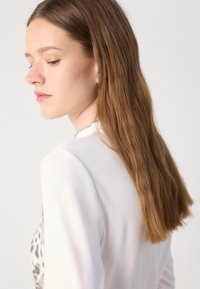 Witte trui met ribbeltextuur, met een gepatterneerd zijontwerp. Lange, steile haren zijn gedeeltelijk zichtbaar. Neutrale achtergrond.