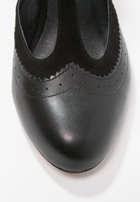 Chaussure en cuir noir avec une finition lisse et des accents en daim en forme de vagues, dotée de perforations décoratives le long du bord et d'un bout arrondi.