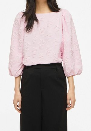 Vrouw draagt een getextureerde lichtroze blouse met 3/4 pofmouwen, gecombineerd met zwarte hoge taille broek tegen een effen achtergrond.