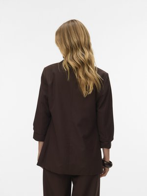 Vrouw met golvend blond haar, gekleed in een donkerbruine blazer en bijpassende broek, staand met de rug naar de camera tegen een effen lichte achtergrond.