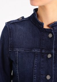 Tmavě modrá džínová bunda s vystouplým límcem, stříbrnými kovovými prvky a předními knoflíkovými zapínáními. Obsahuje dekorativní ramenní švy.