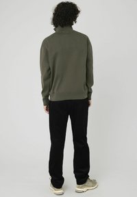Pull en maille vert avec un col montant, des poignets et un ourlet côtelés. Associé à un pantalon en velours côtelé noir et des baskets beiges. Vue de dos.