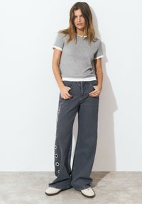 T-shirt gris à manches courtes avec bord blanc ; jeans en denim foncé à jambes larges avec des œillets circulaires sur le côté. Baskets blanches.