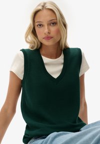 Groene gebreide vest met een v-hals, gedragen over een crèmekleurig t-shirt met korte mouwen, met ribbelranden en een gestructureerde stof. Blauwe jeans zichtbaar.