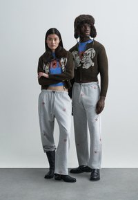 Divi modeļi stāv blakus. Sieviete ir tērpusies īsā brūnā adītā kardigānā ar kažokādu apkakli, zilā krekliņā un pelēkās sporta biksēs. Vīrietis valkā brūnu kardigānu, zilu krekliņu un pelēkas sporta bikses ar sarkaniem lūpu rakstiem, kopā ar melnām zābakām.