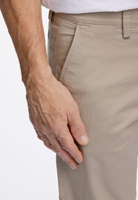 Beige chinos suorat leikkaukset, joissa on hienovaraiset ompeluyksityiskohdat ja sileä kangas. Käsivarsi lepää housun lahkeella.