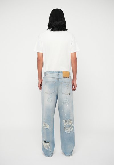 Oseba z dolgimi črnimi lasmi, oblečena v belo majico s kratkimi rokavi in svetlo modre raztrgane kavbojke, stoji obrnjena stran od kamere proti enostavni ozadju.