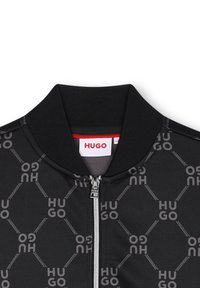 Blouson aviateur noir avec fermeture éclair, arborant un motif répétitif de logo "HUGO" gris et un col côtelé. Texture de tissu lisse.