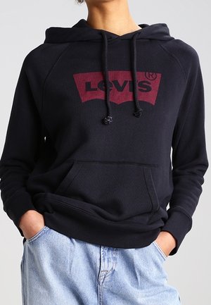 Personne portant un sweat à capuche noir Levi’s avec poche avant et un jean bleu clair, les mains partiellement dans les poches, debout devant un fond uni.