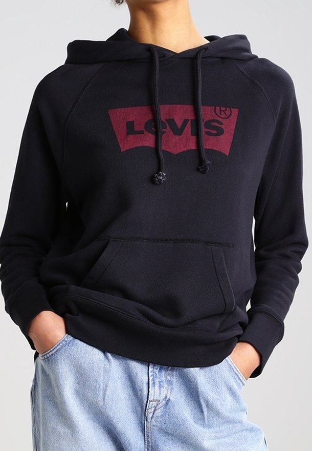 Personne portant un sweat à capuche noir Levi’s avec poche avant et un jean bleu clair, les mains partiellement dans les poches, debout devant un fond uni.