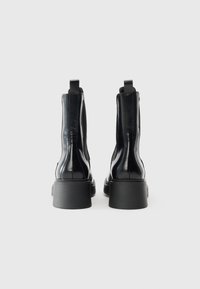 Bottes chelsea noires en cuir avec une finition brillante, panneaux latéraux élastiques, linguets et semelles épaisses noires. Vue de dos mettant en avant la symétrie du design.