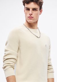 Pánsky krémový rebrovaný sveter s okrúhlym výstrihom, priliehavý strih a malým logom na hrudi. Kombinovaný so strieborným náhrdelníkom.