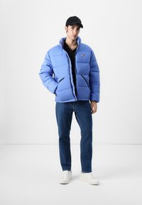 Lyseblå pufferjakke med quiltet tekstur, sorte accenter og et logo-patch. Båret over en sort skjorte med blå jeans og hvide sneakers.