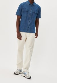 Camicetta di denim a maniche corte blu con due tasche sul petto, chiusura a bottoni e orlo dritto, abbinata a pantaloni color crema e sneakers argentate.