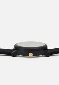 Reloj negro con caja redonda, corona dorada y esfera de cristal lisa. Presenta una correa negra texturizada, que enfatiza un diseño minimalista.