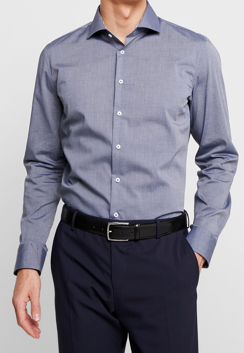 Košile s knoflíky v texturované tmavě modré tkanině, s špičatým límcem, bílými knoflíky a dlouhými rukávy, nošena s černým opaskem.