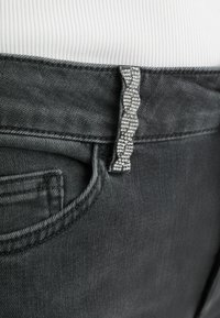 Pelēkie ziloņdāmas džinsi tuvplānā, ar rievotu pelēku auduma detaļu un rhinestone uz jostas. Pastiprināta šuvuma tuvumā pie kabatām.