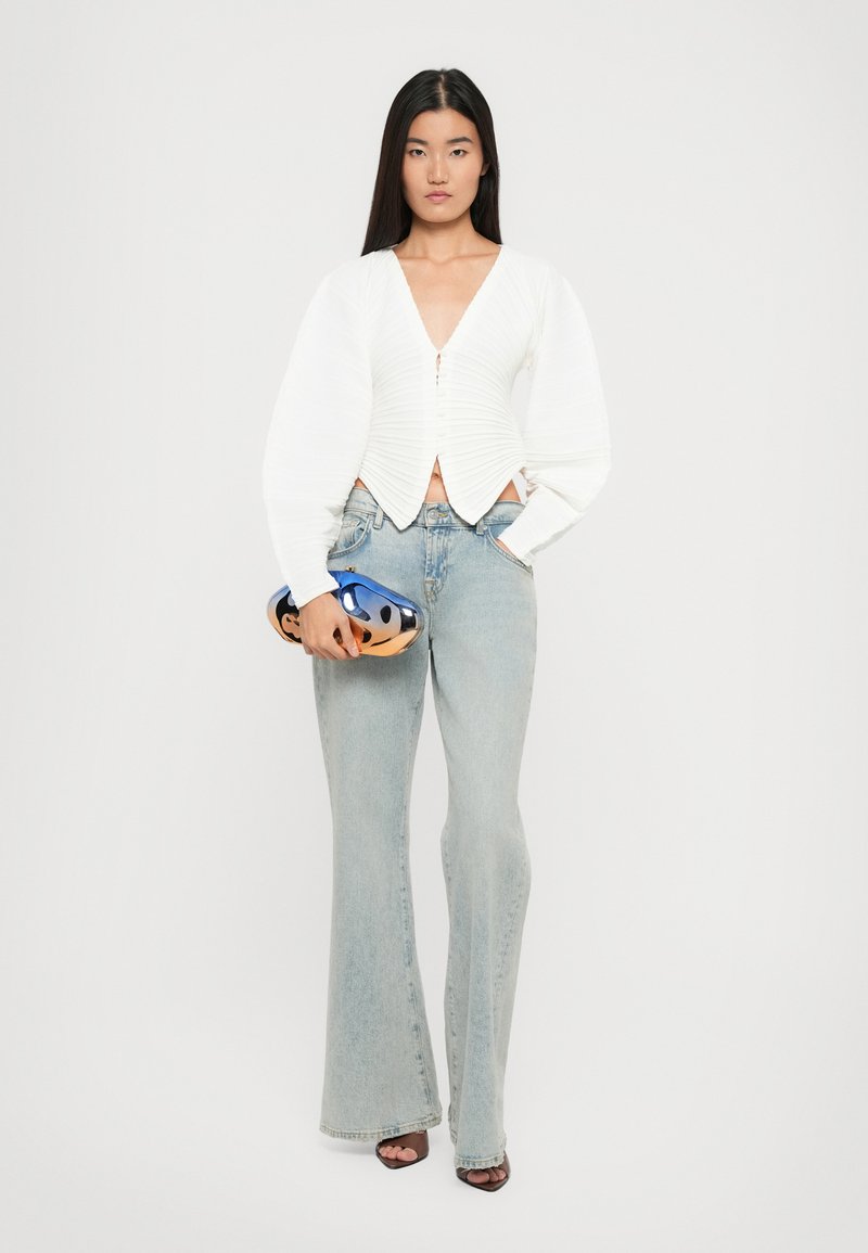 Un haut court texturé blanc avec un décolleté en V plongeant et des boutons, associé à un jean flare bleu clair. Tenant un sac à main métallisé bleu.