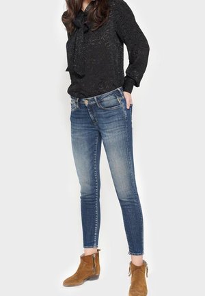 Kobieta ubrana w teksturowaną czarną bluzkę, wyblakłe niebieskie obcisłe jeansy i brązowe zamszowe botki, stojąca na jednolitym tle.