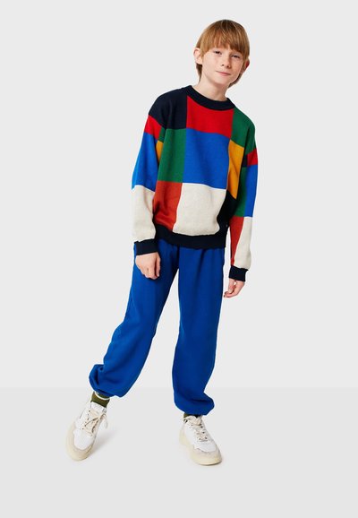 Suéter multicolorido con diseño de patchwork en verde, rojo, azul, amarillo y crema, combinado con pantalones de chándal azules y zapatillas blancas.