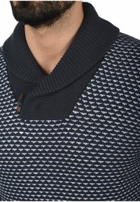 Strikket pullover i marineblå med et tekstureret mønster af hvide zigzag-linjer. Har en bred ribbet sjalskrave og en knapdetalje ved halsen.