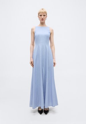 Kvinde med kort blondt hår iført en ærmeløs, gulvlængde lyseblå kjole og sorte spidse sko, stående mod en enkel baggrund.