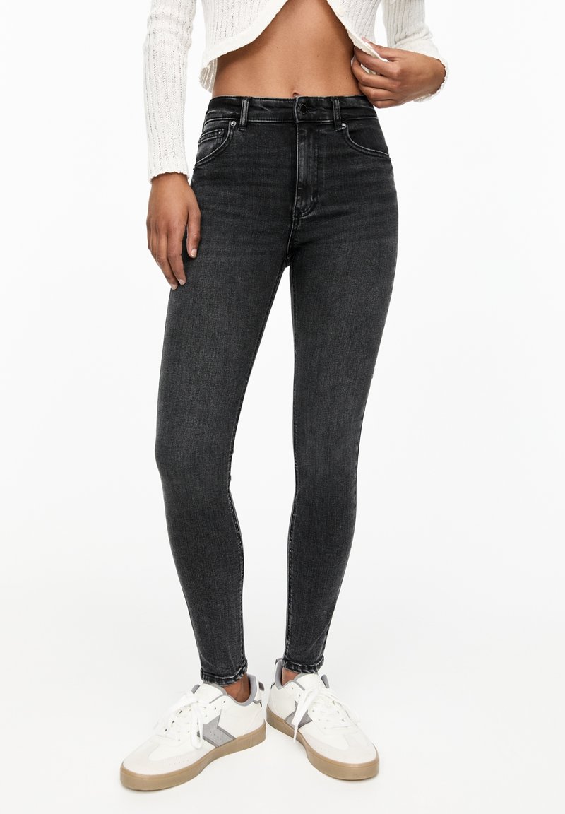 Femme portant un jean skinny taille haute gris foncé, un pull court blanc et des baskets blanches avec des semelles marron clair, debout sur fond blanc.