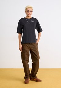 Tricou negru din bumbac cu mâneci scurte și un mic logo alb. Pantaloni maro cu imprimeu leopard verde și adidași maro cu accente portocalii.