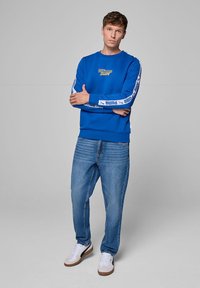 Niebieska bluza z logo "Williams Racing", białe akcenty na rękawach, w połączeniu z jasnoniebieskimi dżinsami i białymi trampkami z brązowymi podeszwami.