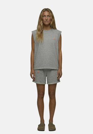 Femme debout face à l'avant portant un haut gris sans manches et un short assorti avec des chaussures slip-on beige sur un fond uni.