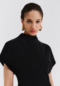 Czarna aksamitna bluzka z wysokim kołnierzem, krótkimi rękawami i miękką fakturą. Charakterystycznym dodatkiem są duże złote kolczyki koła.