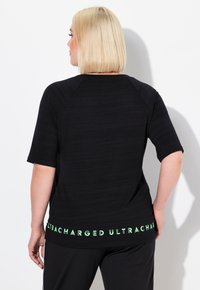 Top negro de manga corta con un patrón texturizado. Presenta una impresión verde de "ULTRACHARGED" en el dobladillo. Ajuste casual con un escote redondeado.