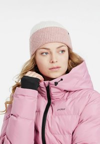 Rosa aufgeplusterte Jacke mit schwarzem Reißverschluss und Logo; gerippte Strickmütze mit weißen und rosa Streifen. Langes, welliges Haar teilweise sichtbar.