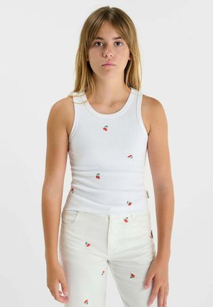 Jeune femme portant un débardeur blanc côtelé et un pantalon blanc, tous deux ornés de petits motifs de cerises rouges, debout devant un fond uni.