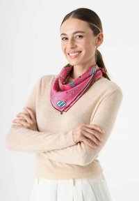 Rosa Seidenschal mit floralen und geometrischen Mustern, akzentuiert mit roten und blauen Streifen. Um den Hals getragen, ergänzt er einen beigen Pullover.