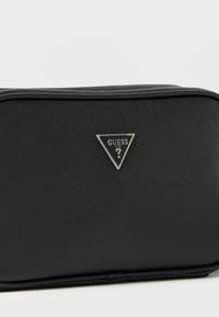 Μαύρη ορθογώνια τσάντα από υλικό με υφή. Διαθέτει ασημί τρίγωνο λογότυπο "GUESS" στο μπροστινό μέρος. Κλεισίματα με φερμουάρ κατά μήκος της επάνω άκρης.