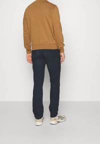 Brun sweatshirt med ribbstickade muddar och fåll, kombinerad med mörkblå strukturerade byxor och beige sneakers med svarta accenter, sedd bakifrån.