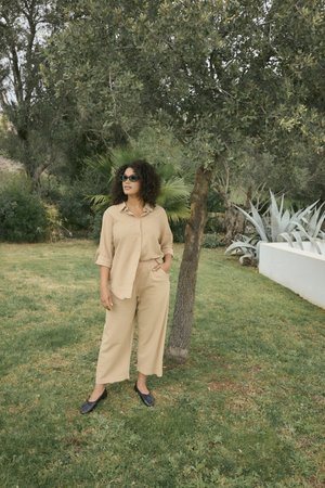 Femme aux cheveux bouclés et lunettes de soleil, debout sur l'herbe à côté d'un arbre, portant une chemise beige et un pantalon avec des ballerines noires dans un cadre de jardin.