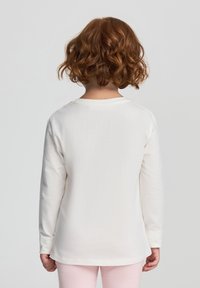 Camicia bianca di cotone a maniche lunghe con scollatura rotonda, tessuto testurizzato e orlo dritto, indossata da un bambino vista di schiena.