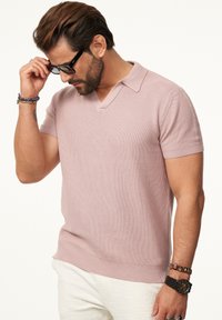 Kurzärmliges, hellrosanes Strickpoloshirt mit V-Ausschnitt und strukturiertem Design, kombiniert mit hellen Hosen und einer schwarzen Uhr.