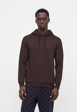 Homme portant un sweat à capuche marron foncé avec une poche avant et des cordons, associé à un pantalon à carreaux bleu foncé, debout devant un fond uni.