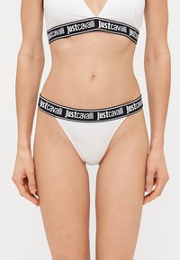 Balts bikini komplekts, kas sastāv no trijstūra braleta un tanga, papildināts ar melnu elastīgo joslu, uz kuras atkārtojoties ir uzraksts "Just Cavalli".