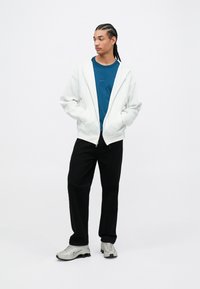 Biely zips sveter, modré tričko s okrúhlym výstrihom, čierne nohavice a šedé športové topánky. Hladká tkanina s voľným strihom a minimalistickým dizajnom.
