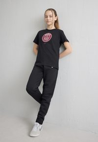 Pusaudze meitene ar pīētām matiem stāv pie balta sienas, valkājot melnu Paris Saint-Germain T-kreklu, melnas bikses un baltus sporta apavus.