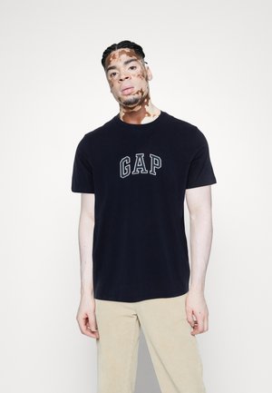 T-shirt en coton bleu marine avec le logo "GAP" en gris clair, col rond, manches courtes, associé à un pantalon en velours côtelé beige.
