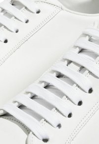 Baskets blanches en cuir avec des lacets plats blancs, des détails cousus et des œillets. Texture lisse et design minimaliste.