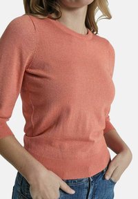 Koraalroze gebreide pullover met driekwart mouwen, ribbelboord en subtiele glans. Getailleerd design dat de taille en mouwen accentueert.
