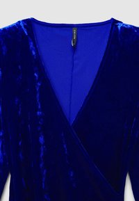 Abito avvolgente in velluto di un blu profondo con scollatura a V. Include un'etichetta appesa, una texture liscia e una superficie lucida che riflette la luce.