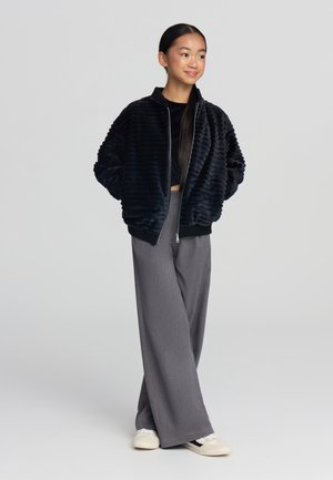 Giacca bomber nera in peluche con texture a coste, chiusura con zip e polsini elasticizzati abbinata a pantaloni grigi larghi e sneakers bianche.