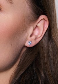 Srebrna kolczyk w kształcie gwiazdy, ozdobiona wieloma małymi niebieskimi kamieniami szlachetnymi, o wypolerowanej powierzchni i bezpiecznym zapięciu widocznym z tyłu.